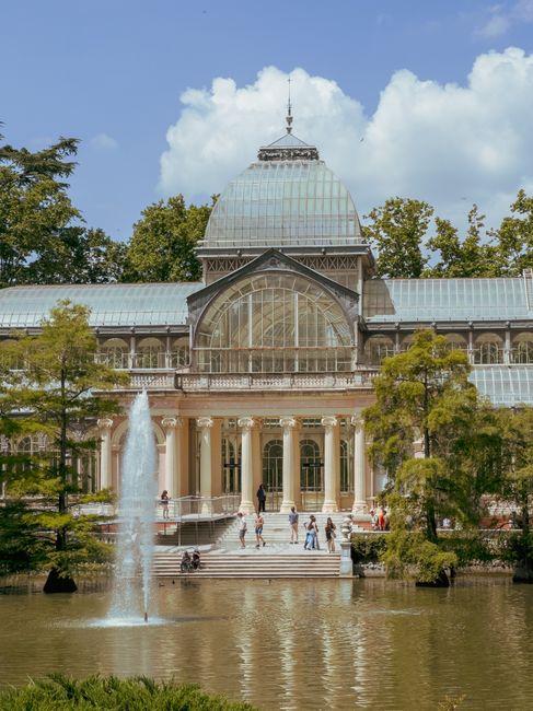 A large historic glass and iron conservatory with a prominent central dome and ornate architecture, situated in a park setting with tall trees and a water feature in the foreground. Several people are