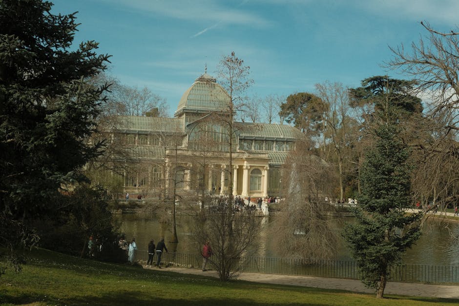 A large historic glass and iron conservatory with a prominent central dome and ornate architecture, situated in a park setting with tall trees and a water feature in the foreground. Several people are