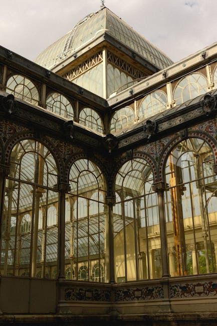 A detailed close-up of the ornate glass and metal framework of a historic greenhouse or conservatory, showing curved glass panes supported by decorative ironwork and metal brackets. The structure's upper section reveals a glass dome with a pyramidal shape, while the lower part displays large, arched windows with intricate patterns and golden accents. The view highlights the conservatory’s architectural features, including the metal trusses and supports, with natural light illuminating the glass panels. This image captures the exterior and interior structural elements typical of vintage glasshouses, relevant to house removals and moving services involving large, fragile structures or historic property furniture transport, such as building disassembly or relocation projects managed by Crystal Palace Movers.