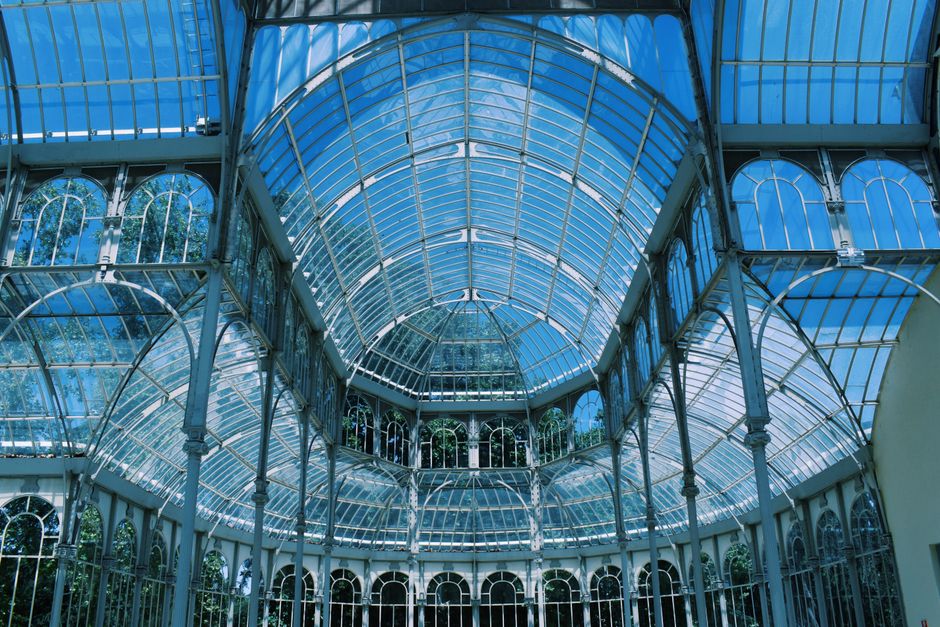 A detailed close-up of the ornate glass and metal framework of a historic greenhouse or conservatory, showing curved glass panes supported by decorative ironwork and metal brackets. The structure's upper section reveals a glass dome with a pyramidal shape, while the lower part displays large, arched windows with intricate patterns and golden accents. The view highlights the conservatory’s architectural features, including the metal trusses and supports, with natural light illuminating the glass panels. This image captures the exterior and interior structural elements typical of vintage glasshouses, relevant to house removals and moving services involving large, fragile structures or historic property furniture transport, such as building disassembly or relocation projects managed by Crystal Palace Movers.