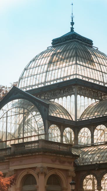 Close-up view of a large, ornate glass and metal conservatory structure with arched windows and decorative metalwork, set against a bright blue sky. The conservatory features multiple levels of curved glass panels supported by intricate metal framework, with visible roof sections and a central dome topped with a small spire. Inside the conservatory, wooden furniture and cardboard packing boxes are visible, indicating ongoing home relocation and packing activities. A moving trolley with straps is positioned near the entrance, and a crystal Palace Movers van is parked outside, near the doorway, contributing to the loading process for house removals in the SE19 Crystal Palace area. The scene captures the detailed architecture and the logistics involved in furniture transport and packing during a same-day move.